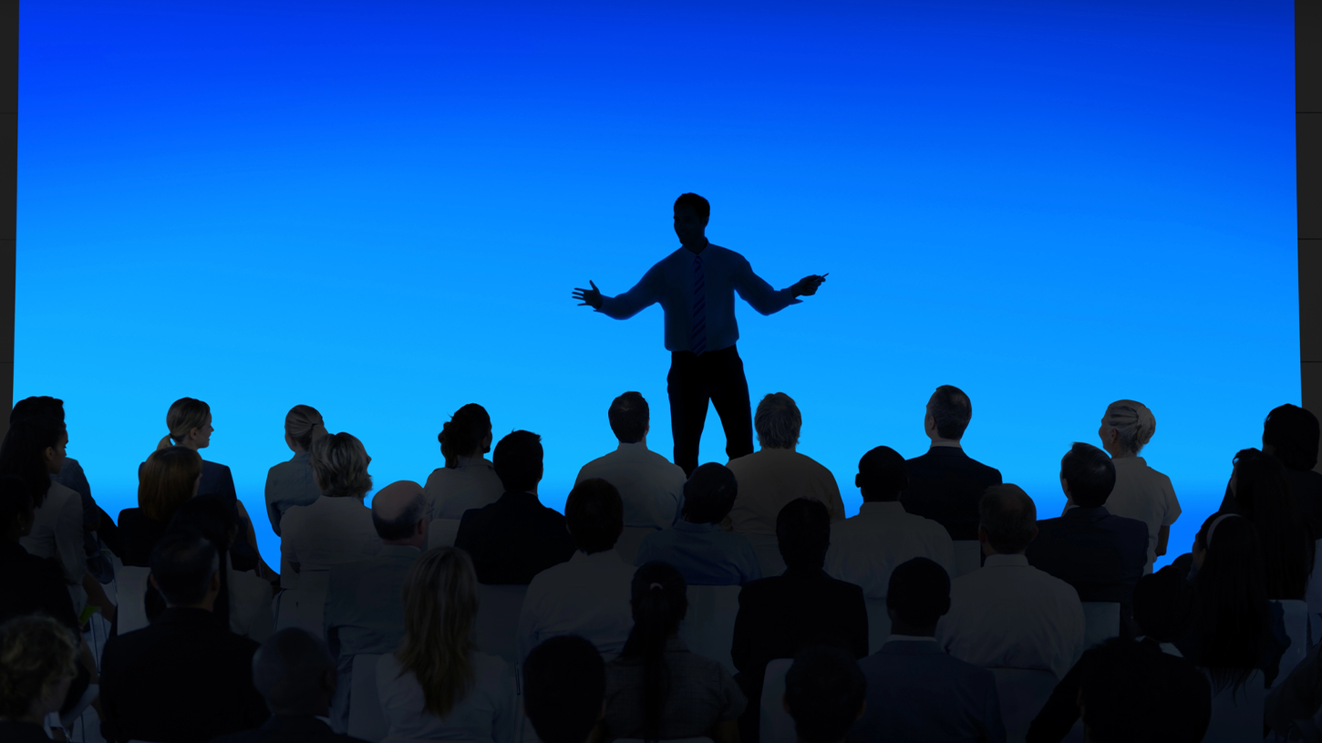 講演中のスピーカーと聴衆、青い背景の講義場にて最新の予防医療と伝統手技について説明。